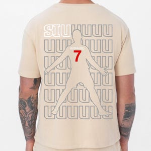 May include: Beige t-shirt featuring a silhouette of a soccer player with outstretched arms. The number 7 is in red, and the name "Ronaldo" is above it. The word "SIUUUU" is at the top in white.
