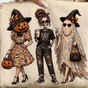 May include: A cream-coloured sweatshirt featuring a graphic of three figures: a person with a pumpkin head wearing a witch hat and dress, a skeleton wearing a t-shirt and jeans holding a coffee cup, and a ghost wearing a witch hat and holding a purse.