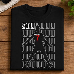 May include: Black crew neck t-shirt featuring a silhouette of a soccer player with outstretched arms. The shirt has the word "SIUUUU" above the player and "Ronaldo 7" in red. The design is repeated in a pattern.