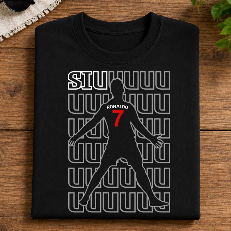 May include: Black crew neck t-shirt featuring a silhouette of a soccer player with outstretched arms. The shirt has the word "SIUUUU" above the player and "Ronaldo 7" in red. The design is repeated in a pattern.
