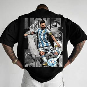 May include: Black t-shirt featuring a graphic of a soccer player in a blue and white striped jersey kicking a soccer ball. The player's name, "Lionel Messi," is printed in large white letters. The shirt has a crew neck and short sleeves.