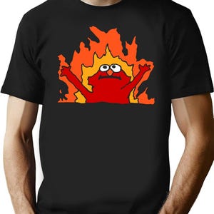 May include: Black t-shirt featuring a distressed graphic of a red cartoon character engulfed in flames. The character has wide eyes and raised arms, set against a backdrop of orange and yellow flames. The shirt is a classic crew neck.