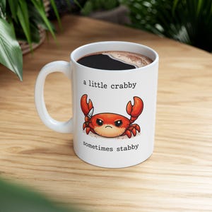 May include: White ceramic mug with a black coffee fill. The mug features a cartoon crab illustration with the text "a little crabby sometimes stabby". The crab is red with orange claws and a stern expression.