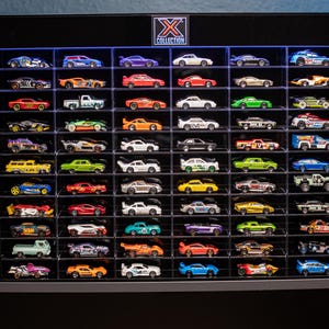 May include: A black display case filled with miniature toy cars of various colors and designs. The case has multiple shelves, each holding several cars. The top of the case features the text "X COLLECTION" and a logo. The cars are arranged neatly.