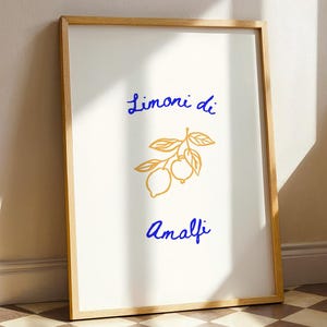May include: A framed art print featuring the words "Limoni di Amalfi" in blue script, with a gold illustration of lemons and leaves. The artwork has a light wood frame and is leaning against a wall.