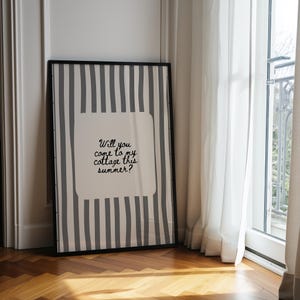 May include: Framed art print with a black frame. The artwork features a white background with a gray striped pattern and a central white square containing the text: "Will you come to my cottage this summer?" The print is leaning against a wall.