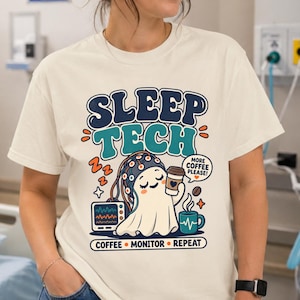 Peut inclure: Un t-shirt couleur crème avec un motif représentant un fantôme portant un bonnet EEG, tenant une tasse de café et assis à côté d'un moniteur médical. Le texte indique SLEEP TECH, MORE COFFEE PLEASE et COFFEE MONITOR REPEAT en lettres colorées.
