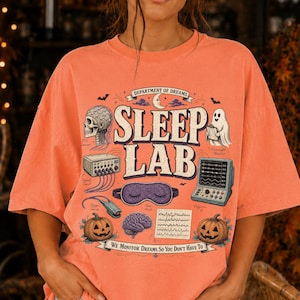 Könnte beinhalten: Ein orangefarbenes T-Shirt mit einem Grafikdruck, der den Text "Department of Dreams Sleep Lab" und "We monitor dreams so you don't have to" enthält. Das Design zeigt Illustrationen eines Schädels, eines Geistes, Kürbissen, eines Gehirns und medizinischer Geräte.