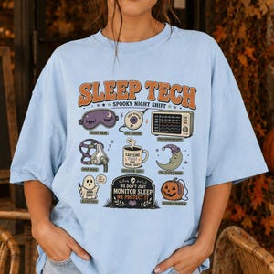 May include: A light blue t-shirt featuring a graphic with the text SLEEP TECH SPOOKY NIGHT SHIFT. The design includes illustrations of a sleep mask, sensor, monitor, CPAP mask, coffee mug, moon, ghost, tombstone, and pumpkin with the text WE DON'T JUST MONITOR SLEEP WE PROTECT IT.
