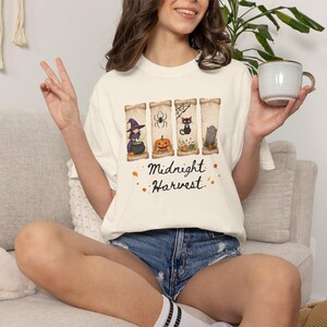 Puede incluir: Camiseta color crema con un diseño temático de Halloween. El diseño presenta una bruja, una araña, un gato negro y una lápida. Las palabras "Midnight Harvest" están impresas debajo. La camiseta se usa con pantalones cortos de mezclilla.