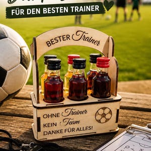 Puede incluir: Un portabotellas de madera que contiene seis botellas de vidrio en miniatura con tapas rojas y negras. El soporte presenta un grabado láser con el texto "BESTER Trainer" en el asa y "OHNE Trainer KEIN Team DANKE FÜR ALLES!" en la parte frontal.