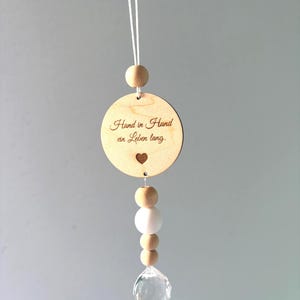 Könnte beinhalten: Ein Holzornament mit dem Text "Hand in Hand an Leben lang" und einem kleinen Herz. Das runde Ornament ist an einer Schnur aufgehängt. Darunter befinden sich Holz- und weiße Perlen sowie ein klarer Kristallkugel.
