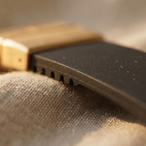 May include: Close-up of a watch band with a black rubber strap and a gold-colored metal clasp. The strap has a textured surface and is partially visible against a neutral fabric background.