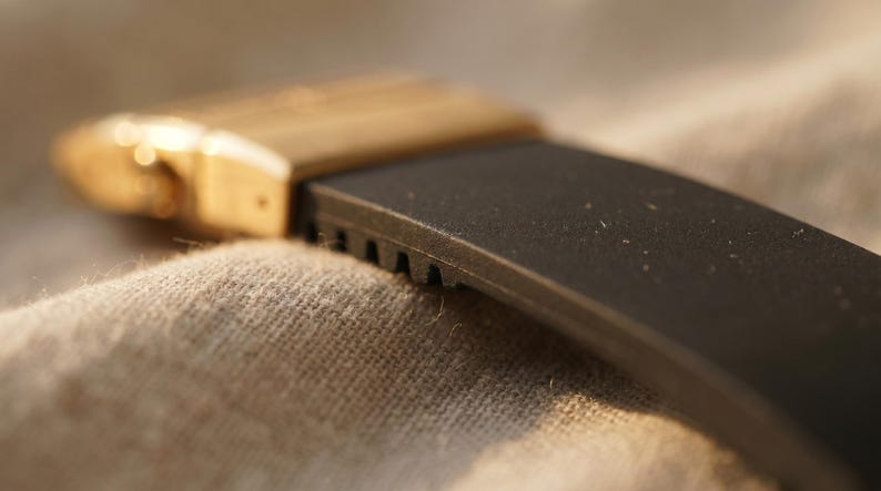May include: Close-up of a watch band with a black rubber strap and a gold-colored metal clasp. The strap has a textured surface and is partially visible against a neutral fabric background.