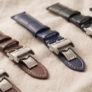 May include: Close-up of several leather watch straps in various colors, including brown, navy blue, and black. Each strap features a stainless steel clasp and is displayed on a neutral-toned fabric surface.