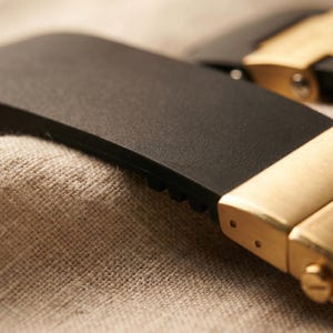 May include: Close-up of a black watch strap with a gold-colored clasp. The clasp is rectangular with the brand name "ULYSSE NARDIN" engraved on it. The strap is made of a flexible material and is resting on a textured, beige surface.