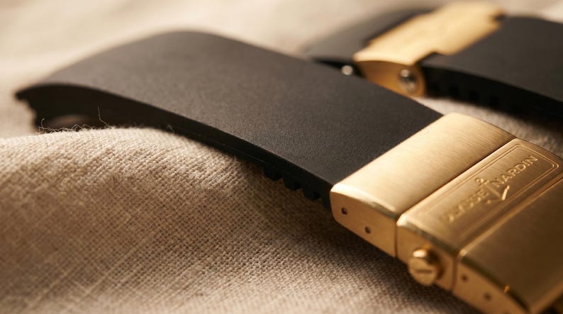 May include: Close-up of a black watch strap with a gold-colored clasp. The clasp is rectangular with the brand name "ULYSSE NARDIN" engraved on it. The strap is made of a flexible material and is resting on a textured, beige surface.