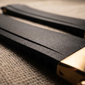 May include: Close-up of two black watch straps with gold-colored clasps. The straps have a textured surface and are angled towards the viewer. The clasps are engraved with the brand name "Ulysse Nardin".