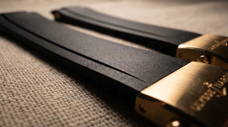 May include: Close-up of two black watch straps with gold-colored clasps. The straps have a textured surface and are angled towards the viewer. The clasps are engraved with the brand name "Ulysse Nardin".