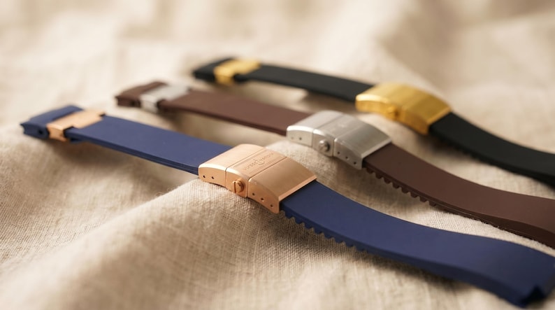 May include: Close-up of four watch straps in various colors: navy blue, brown, and black. Each strap has a different metal clasp in rose gold, silver, and gold. The straps are made of a flexible material.