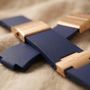 May include: Close-up of a navy blue watch strap with rose gold clasps. The strap is made of a flexible material and features a rectangular design. The clasps have a polished finish and are engraved with a brand name. The watch strap is displayed on a textured, beige surface.