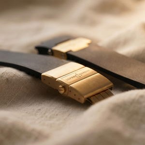 May include: Two black watch straps with gold-colored clasps, one in focus, the other slightly blurred in the background. The clasps are rectangular with the text "ULYSSE NARDIN" engraved. The straps rest on a beige, textured surface.