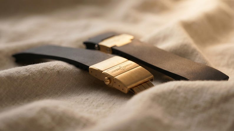 May include: Two black watch straps with gold-colored clasps, one in focus, the other slightly blurred in the background. The clasps are rectangular with the text "ULYSSE NARDIN" engraved. The straps rest on a beige, textured surface.