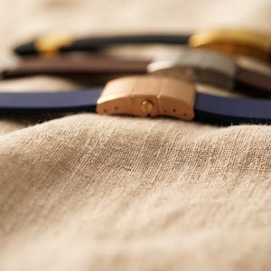 May include: Close-up of several watch straps in various colors, including navy blue, brown, and black. The straps have metal clasps in gold, silver, and rose gold tones. The straps are resting on a textured, beige fabric.