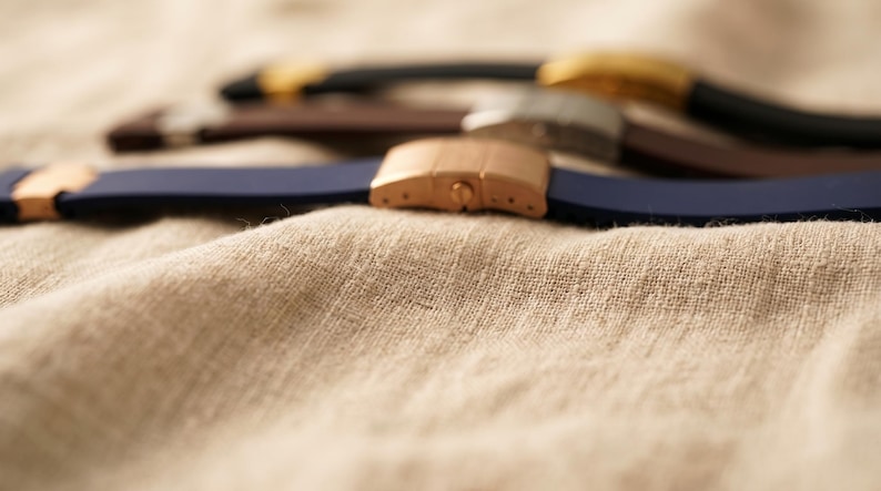 May include: Close-up of several watch straps in various colors, including navy blue, brown, and black. The straps have metal clasps in gold, silver, and rose gold tones. The straps are resting on a textured, beige fabric.