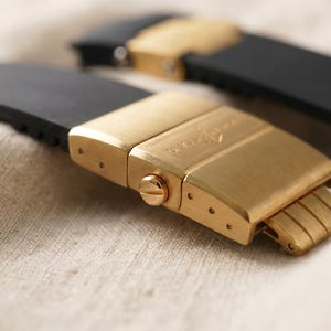 May include: Close-up of two black watch straps with gold-colored clasps. The clasps are rectangular with a brushed finish and a small screw detail. The straps are made of a flexible material, likely rubber or silicone.