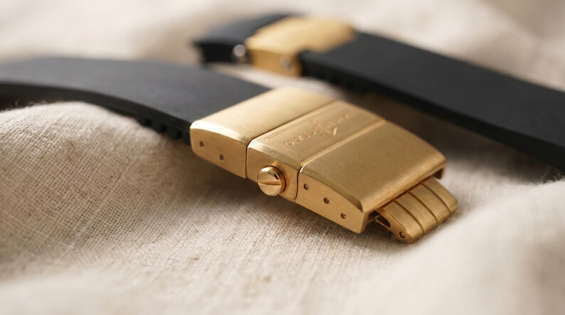 May include: Close-up of two black watch straps with gold-colored clasps. The clasps are rectangular with a brushed finish and a small screw detail. The straps are made of a flexible material, likely rubber or silicone.