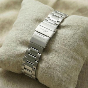 May include: A silver-toned wristwatch with a metal band rests on a small, light-colored pillow. The watch clasp is visible, displaying the brand name and logo. The watch band is made of multiple rectangular links.