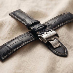 May include: A black leather watch strap with a crocodile pattern. The strap features a silver-colored clasp with the brand name "Longines" engraved on it. The strap is lying on a beige fabric.