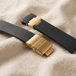 May include: Two black watch straps with gold-colored clasps are displayed on a beige fabric. The clasps have a rectangular shape with a logo. The straps are made of a flexible material.
