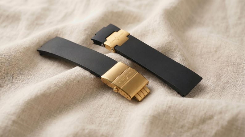 May include: Two black watch straps with gold-colored clasps are displayed on a beige fabric. The clasps have a rectangular shape with a logo. The straps are made of a flexible material.