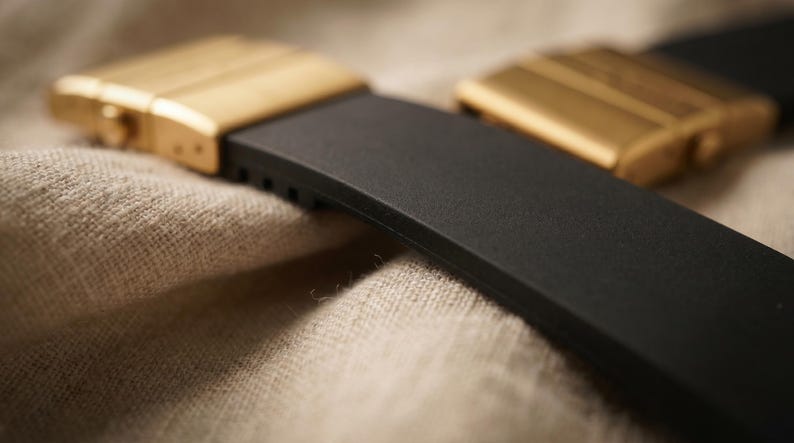 May include: Close-up of a black watch strap with gold-colored metal clasps. The strap is made of a flexible material, and the clasps have a rectangular design with horizontal lines. The background is a textured, neutral-colored fabric.
