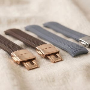 Puede incluir: Dos correas de reloj de silicona marrón con cierres desplegables de metal en tono oro rosa y dos correas de silicona gris con cierres desplegables de metal en tono plateado, todas con un patrón de rejilla texturizado en la superficie.