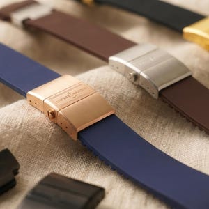 May include: Close-up of several watch straps in various colors, including navy blue, brown, and black. The straps have metal clasps in gold, silver, and rose gold tones. The straps are made of a flexible material, likely silicone or rubber.