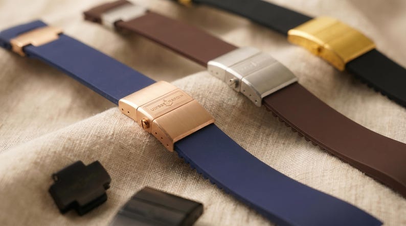 May include: Close-up of several watch straps in various colors, including navy blue, brown, and black. The straps have metal clasps in gold, silver, and rose gold tones. The straps are made of a flexible material, likely silicone or rubber.