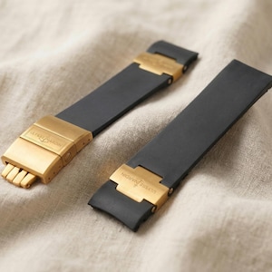 May include: Two black watch straps with gold-coloured clasps. The clasps have a rectangular design with the brand name engraved. The straps are made of a flexible material and are displayed on a beige fabric surface.