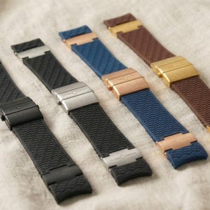 May include: Four watch straps in black, blue, and brown, with metal clasps in black, silver, rose gold, and gold. The straps have a textured surface and are arranged diagonally on a neutral fabric background.
