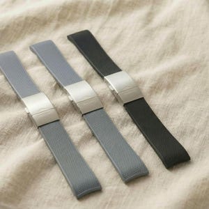 May include: Four watch straps in a row, two gray and one black, with silver clasps. The straps are made of a textured material and are laid out on a beige fabric background.