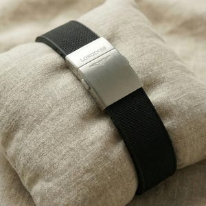 May include: A black watch strap with a silver clasp, featuring the brand name "LONGINES", rests on a small, beige pillow. The strap has a textured surface. The pillow is made of a light-colored, textured fabric.
