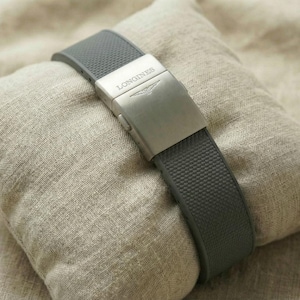 May include: A gray watch band with a textured surface and a silver clasp, resting on a small, beige pillow. The clasp is engraved with the brand name "LONGINES" and a logo. The watch band is made of a flexible material.