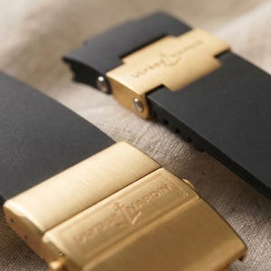 May include: Close-up of two black watch straps with gold-colored clasps. The clasps are rectangular and feature the brand name. The straps are made of a smooth, matte material. The background is a neutral, textured fabric.