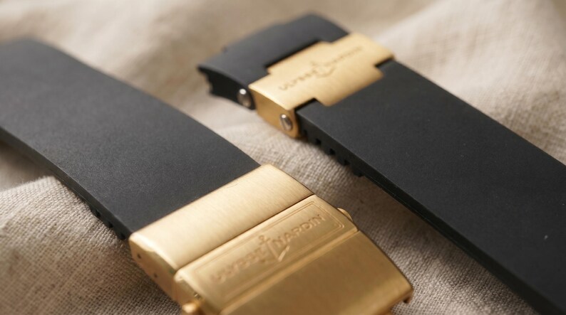 May include: Close-up of two black watch straps with gold-colored clasps. The clasps are rectangular and feature the brand name. The straps are made of a smooth, matte material. The background is a neutral, textured fabric.