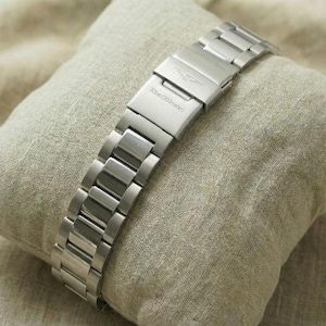 May include: A silver-toned watch band with a clasp, resting on a small, square, beige pillow. The watch band is made of rectangular links. The pillow is on a beige, textured surface.