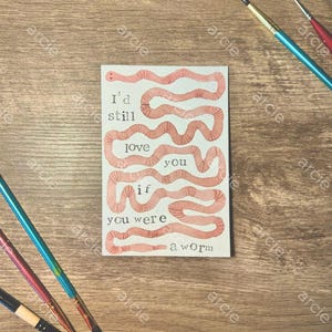 May include: A rectangular art print with the text "I'd still love you if you were a worm" in black font. The print features a pink worm design. Paintbrushes in blue, red, and black are scattered around the print on a wooden surface.
