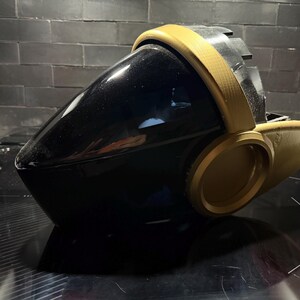Puede incluir: Un casco futurista con una visera negra brillante y lisa que se curva hacia una punta frontal. La estructura está enmarcada por un borde de color dorado mate que rodea la visera y se extiende hacia paneles laterales circulares texturizados.