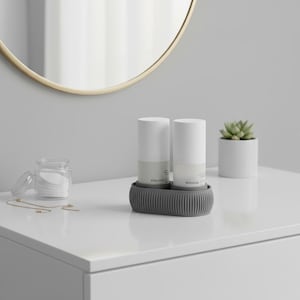 Puede incluir: Una escena de baño minimalista con dos botellas cilíndricas blancas en un soporte gris. También hay una pequeña suculenta en una maceta blanca y un tarro de discos de algodón. Un espejo con marco dorado cuelga sobre la superficie blanca.